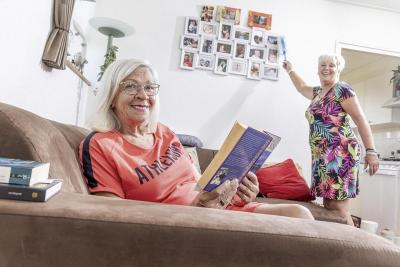 Thuishulp Els aan het stoffen en klant kijkt vanaf bank lachend toe. Thuishulp Els aan het stoffen en klant kijkt vanaf bank lachend toe.